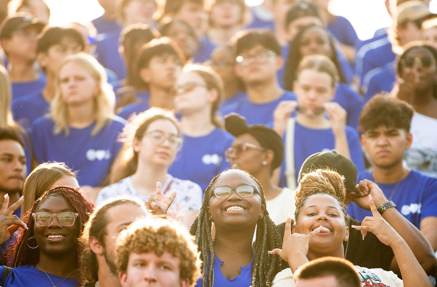 A diverse group of young people in blue shirts cheer and gesture excitedly at an outdoor event under bright sunlight.